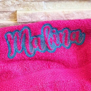 May include: A bright pink towel with the name "Malina" embroidered in a cursive font. The name is outlined in teal and filled with a textured, light blue fabric.