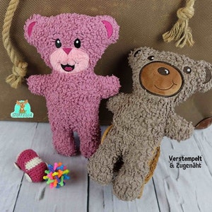 May include: Two plush teddy bears, one pink and one brown, with embroidered faces. The pink bear has a happy expression and the brown bear has a neutral expression. The bears are sitting on a wooden surface. The text "Verstempelt & Zugenaeht" is visible in the lower right corner of the image.