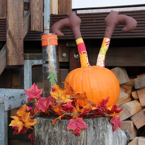 May include: A pumpkin with fabric legs sticking out of the top, decorated with colorful patterns. The pumpkin is surrounded by fall leaves and is sitting on a tree stump.