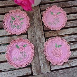 Könnte beinhalten: Vier rosa gehäkelte Untersetzer mit gestickten Rosen und grünen Blättern. Die Untersetzer liegen auf einer Holzoberfläche.