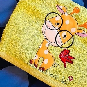 Könnte beinhalten: Ein gelbes Handtuch mit einer gestickten Giraffe, die eine Brille trägt und einen roten Vogel. Die Giraffe hat weiße Flecken und ein lächelndes Gesicht.