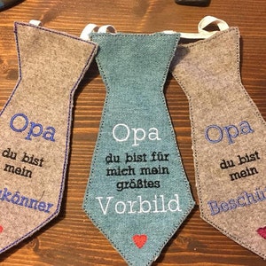 Könnte beinhalten: Drei graue Filz-Krawatten-Anhänger mit blauer Stickerei. Jede Krawatte trägt das Wort "Opa" und eine andere Botschaft: "du bist mein Alleskönner", "du bist für mich mein größtes Vorbild" und "du bist mein Beschützer".