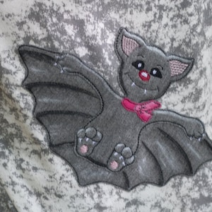 May include: A gray embroidered bat with pink accents, including a bow tie and feet, is flying with its wings spread out.