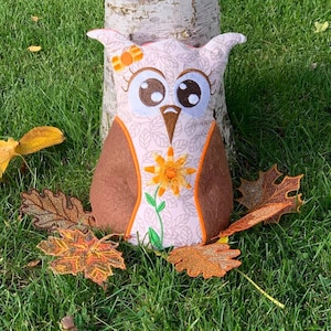 May include: A plush owl toy with brown and white fabric, large eyes, and a flower on its head. The owl is sitting on a bed of green grass with orange and brown leaves.