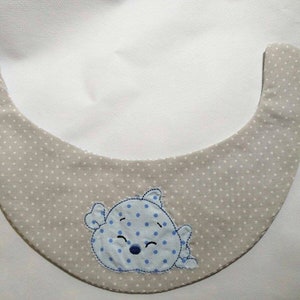 May include: A beige baby bib with white polka dots and a blue and white embroidered cartoon animal design.