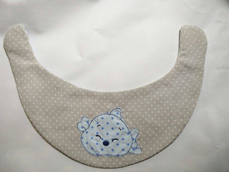 May include: A beige baby bib with white polka dots and a blue and white embroidered cartoon animal design.
