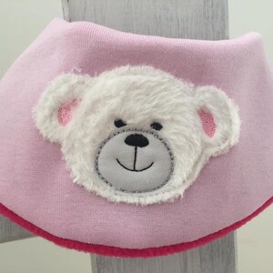 May include: Pink baby bib with a white fuzzy bear face applique. The bear has a gray nose and mouth.