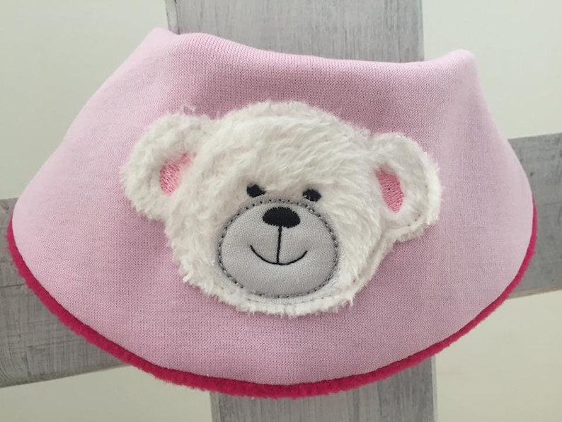 May include: Pink baby bib with a white fuzzy bear face applique. The bear has a gray nose and mouth.