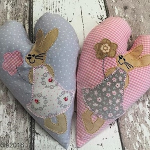 Könnte beinhalten: Zwei Stoffkissen in Herzform mit gestickten Hasendesigns. Ein Kissen ist rosa mit weißem Gingham und das andere ist grau mit weißen Punkten.