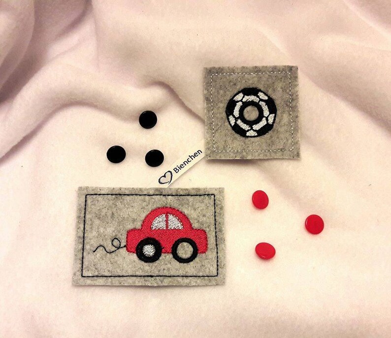 May include: Three felt patches with embroidered designs. One patch features a red car with a white windshield and wheels. Another patch features a black and white circle design. The third patch has the word "Bienchen" in white with a heart symbol.