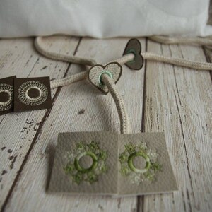 以下が含まれることがあります： 白い刺繍の円が施された茶色の布地正方形2つ、白い刺繍の円と緑色のアクセントが施された茶色の布地ハート2つ、緑色の刺繍の円が施されたベージュの布地正方形2つ。布地は白いロープで繋がっている。