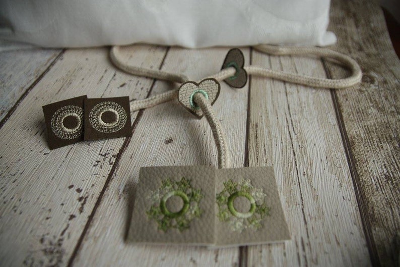以下が含まれることがあります： 白い刺繍の円が施された茶色の布地正方形2つ、白い刺繍の円と緑色のアクセントが施された茶色の布地ハート2つ、緑色の刺繍の円が施されたベージュの布地正方形2つ。布地は白いロープで繋がっている。