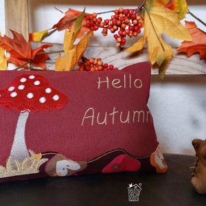 May include: A red and white mushroom embroidered on a burgundy pillow with the text "Hello Autumn". The pillow is sitting on a wooden shelf with fall foliage and a brown hedgehog figurine.
