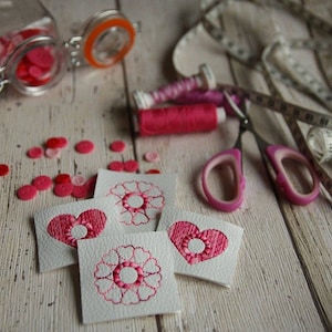 以下が含まれることがあります： ピンクの刺繍が施されたハートと花がついた白いフェルトの正方形が4つ。ピンクのボタン、ピンクの糸巻き、ピンクのハサミ、メジャーが木製の表面に置かれています。
