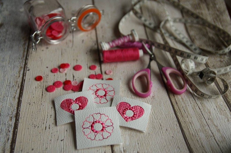 以下が含まれることがあります： ピンクの刺繍が施されたハートと花がついた白いフェルトの正方形が4つ。ピンクのボタン、ピンクの糸巻き、ピンクのハサミ、メジャーが木製の表面に置かれています。