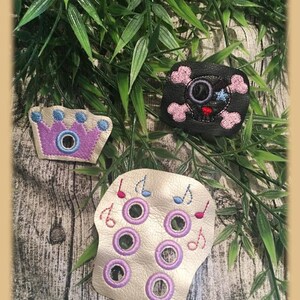 May include: Three felt patches with embroidered designs. A purple crown with blue accents, a black skull with pink accents, and a gold skull with purple accents and musical notes.