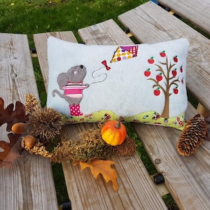 May include: A grey and pink mouse holding a kite flies near an apple tree with red apples. The image is on a white pillow with a green and white border. The pillow is on a wooden bench with autumn leaves and acorns.
