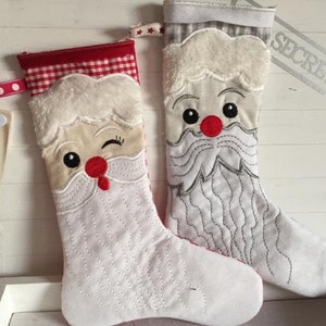 Könnte beinhalten: Zwei weiße Weihnachtssocken mit gestickten Weihnachtsmann-Gesichtern. Ein Socken hat einen roten und weißen karierten Bund und der andere einen grauen und weißen karierten Bund. Der Socken mit dem roten und weißen karierten Bund hat ein zwinkerndes Weihnachtsmann-Gesicht. Der Socken mit dem grauen und weißen karierten Bund hat ein ernstes Weihnachtsmann-Gesicht.