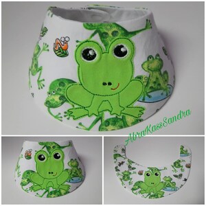 May include: White fabric baby bib with a green frog design. The bib has a green frog with big eyes and a small smile in the center. The bib is surrounded by smaller green frogs with white spots.