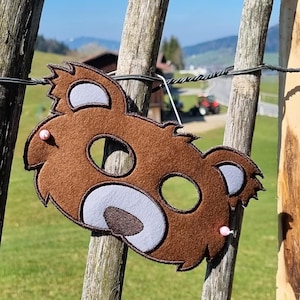 Puede incluir: Una máscara de oso de fieltro marrón con nariz y orejas de color azul claro. La máscara está colgada en una valla de madera.