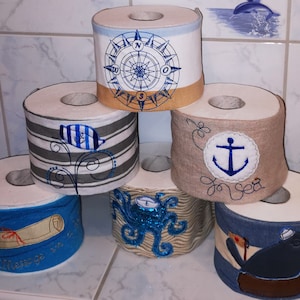 Pode incluir: Seis rolos de papel higiênico decorados com temas náuticos. Os rolos apresentam desenhos como uma bússola, uma âncora, um polvo e uma mensagem em uma garrafa.