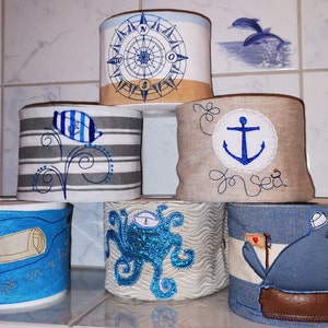 Könnte beinhalten: Sechs dekorierte Toilettenpapierrollen mit nautischen Motiven. Die Rollen zeigen einen Kompass, einen Anker, einen Oktopus, einen Vogel und eine Schriftrolle. Die Rollen sind aus Stoff und haben verschiedene Farben und Muster.