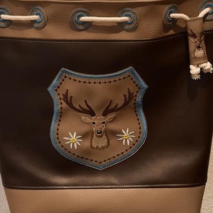 May include: A brown and tan leather bucket bag with a drawstring closure. The bag features an embroidered shield with a deer head and floral accents. The top has a white rope drawstring and blue accents.