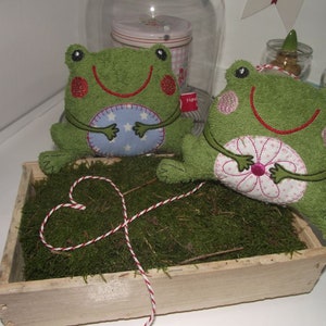 May include: Two green frog plushies with embroidered details, sitting in a wooden box filled with moss. One frog holds a blue and white circle, the other holds a pink and white flower.