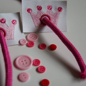 May include: Two pink felt crowns with pink thread embroidery and pink buttons on a white surface. A pink cord is threaded through the crowns.