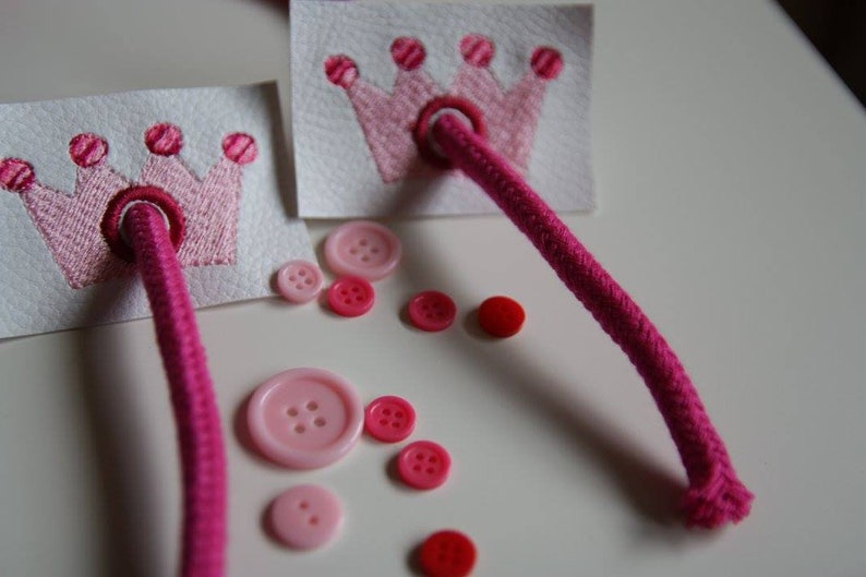 May include: Two pink felt crowns with pink thread embroidery and pink buttons on a white surface. A pink cord is threaded through the crowns.