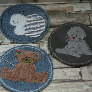 May include: Three round fabric patches with embroidered designs. One features a light blue snail with a striped shell, another a gray seal, and the third a brown teddy bear. The patches are on a denim background, with a needle and scissors visible.