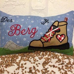 May include: A blue pillow with white snowflakes and the text "Der Berg ruft" embroidered in red. A brown hiking boot with red and white laces and a red and white checkered heart is embroidered on the pillow.