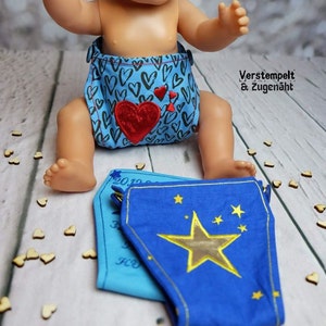May include: A blue and white nappy with a red heart and the text "Verstempelt & Zugenaeht" on a doll. The doll is wearing the nappy and has a blue and gold star patterned nappy on top of the nappy.
