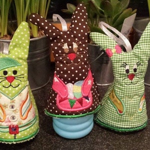 Może przedstawiać: Trzy wielkanocne dekoracje w kształcie królików. Jeden jest zielony w białe kropki, jeden brązowy w białe kropki, a jeden zielony w kratkę. Każdy królik ma inną haftowaną twarz i strój. Mają około 15 cm wysokości.