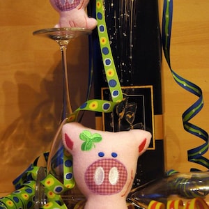 May include: Two pink pig plush toys with embroidered details, one perched on a wine glass. A black bottle is in the background, adorned with colorful ribbons and festive party streamers. The pig's snout has a checkered pattern.