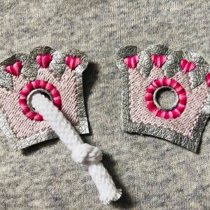 May include: Two silver and pink crown-shaped fabric patches with pink stitching and a hole in the center. The patches are attached to a white cord.