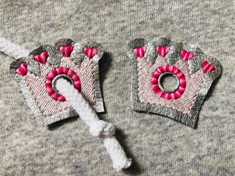 May include: Two silver and pink crown-shaped fabric patches with pink stitching and a hole in the center. The patches are attached to a white cord.