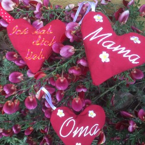 May include: Three red felt heart-shaped ornaments with white embroidery. The hearts are decorated with white flowers and the words "Ich hab dich lieb", "Mama", and "Oma".