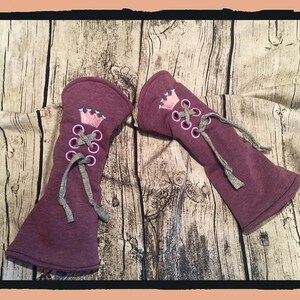 May include: A pair of maroon fabric arm warmers with a pink crown and gray laces. The arm warmers are on a wooden background.