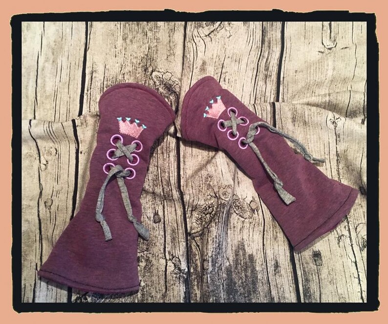 May include: A pair of maroon fabric arm warmers with a pink crown and gray laces. The arm warmers are on a wooden background.