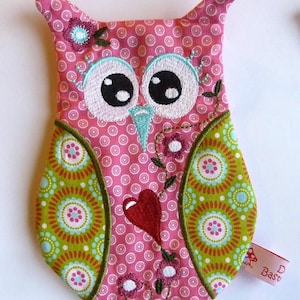 Könnte beinhalten: Rosa geformtes Stofftier in Form einer Eule mit gestickten Augen, einem Herzen und floralen Details. Der Stoff hat ein Punkte-Muster und ein grünes und rotes Kreis-Muster.