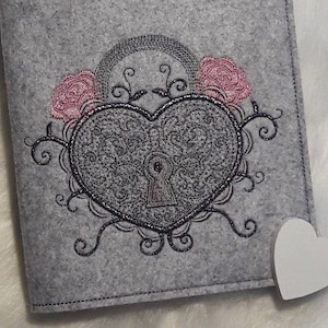 Könnte beinhalten: Ein grauer Filz-Notizbuchumschlag mit einem gestickten Design eines herzförmigen Schlosses mit einem Schlüsselloch, umgeben von rosa Rosen und wirbelnden Ranken.