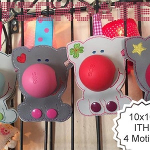 Könnte beinhalten: Vier EOS-Lippenbalsamhalter in Bärenform, jeder mit einem andersfarbigen Balsam. Die Halter sind weiß und grau mit gestickten Details und an Schlüsselanhängern mit bunten Bändern befestigt. Der Text "10x10 ITH 4 Motive" befindet sich unten rechts.