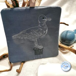 Könnte beinhalten: Eine blaue Stofftasche mit einem weißen gestickten Bild einer Möwe, die auf einem Pfosten steht. Die Tasche hat einen weißen Henkel.