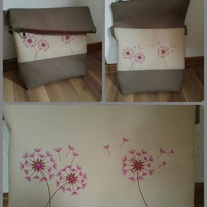 Op de afbeelding: Een beige en bruine stoffen tas met een ritssluiting en een ontwerp van roze paardenbloemen met zaden die in de wind waaien.