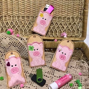 Puede incluir: Cuatro bolsas de tela en forma de cerdo rosa con detalles bordados, cada una con una ranura para una unidad USB. Las bolsas se muestran en una cesta de mimbre con un bálsamo labial rosa y una unidad USB verde.