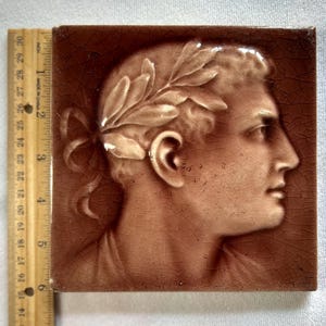 May include: Square brown tile featuring a profile of a person with a laurel wreath. The relief image is in a lighter shade of brown, with a crackled texture. The tile is approximately 4 inches square.