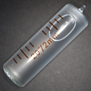 May include: A clear glass graduated cylinder with a brown printed scale marked in 2g/2ml increments. The cylinder has a rounded bottom and a small opening at the top.