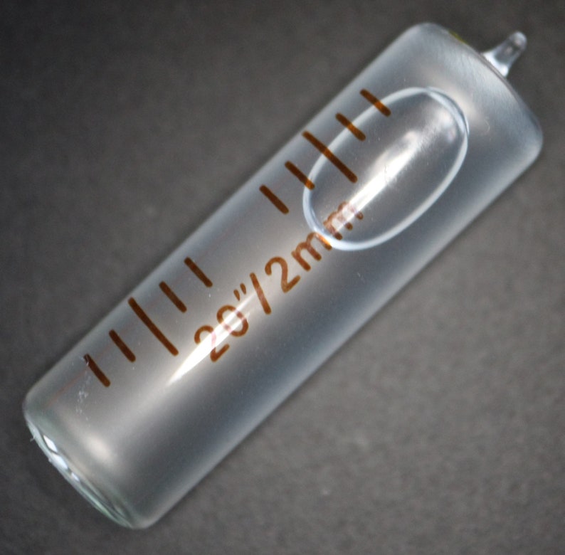 May include: A clear glass graduated cylinder with a brown printed scale marked in 2g/2ml increments. The cylinder has a rounded bottom and a small opening at the top.