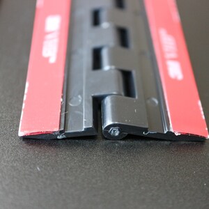 Op de afbeelding: Close-up van een zwart plastic scharnier met rode plakband aan beide zijden. Het scharnier is gemaakt van meerdere delen die in elkaar passen.